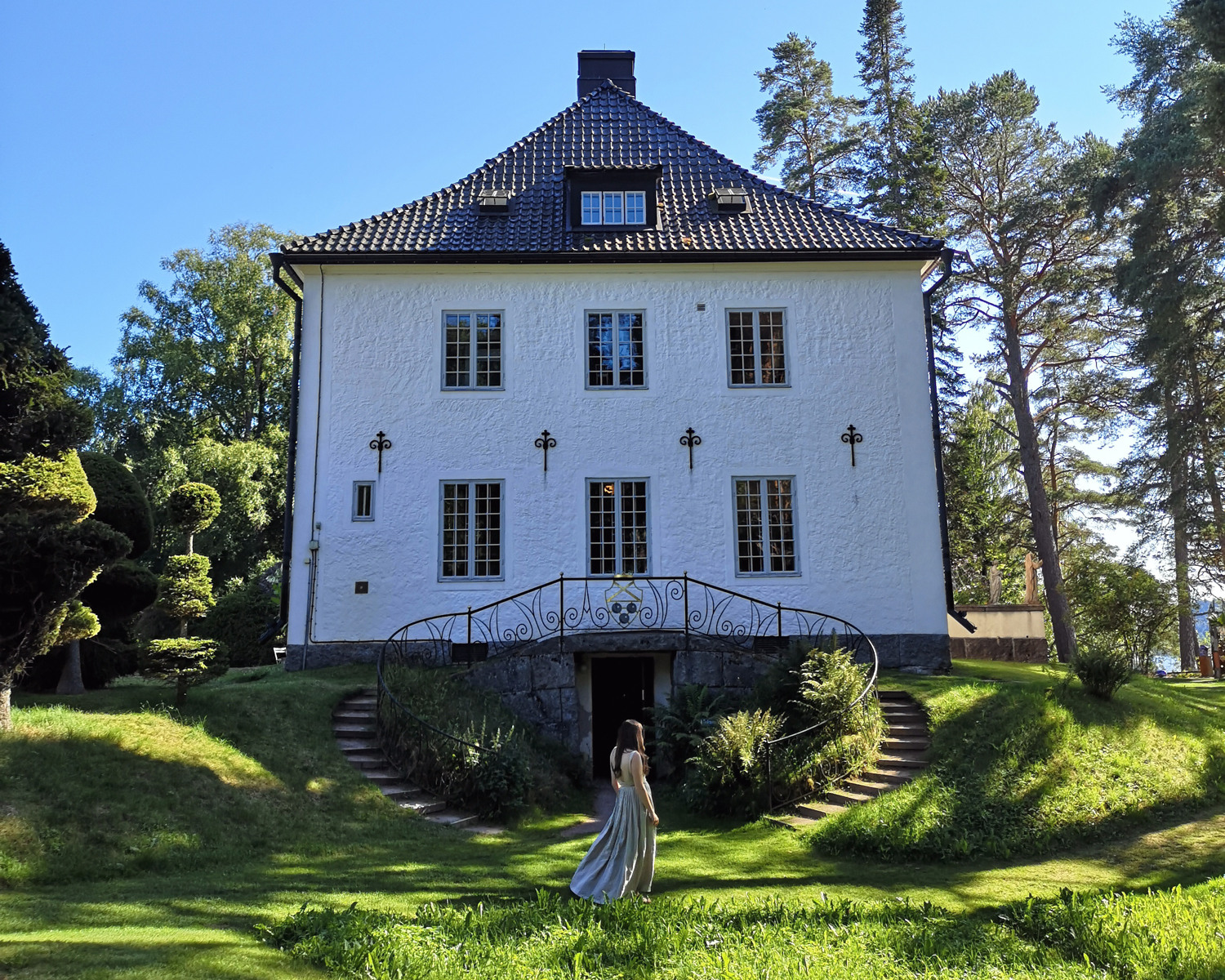 En kvinna går i en trädgård framför ett vitt hus.