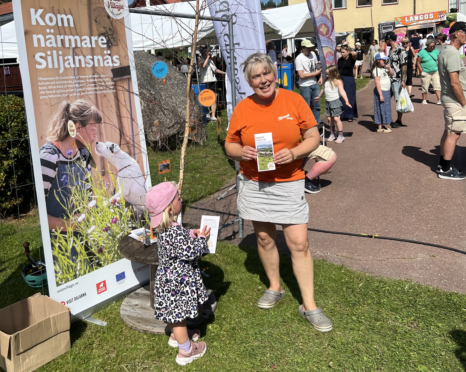 En kvinna står på en mässa utomhus intill en skylt.