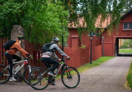 Två personer cyklar sommartid bland faluröda hus.