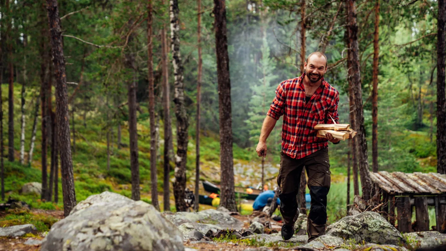 Man i rödrutig skjorta bär ved genom skog.
