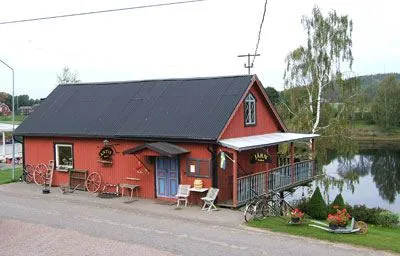 Fäggeby Antikbod Ett rött hus med en blå dörr, brevid dörren två stolar och ett bord, huset ligger precis brevid vattnet.