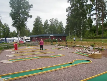 En bangolfbana, två personer som spelar, en brun låg byggnad och husvagnar i bakgrunden.