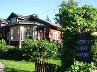 En röd träskylt med texten antikt, konst, inredning, en trädgård och ett rött trähus i bakgrunden.