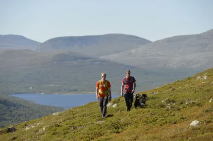 Två vandrare med hund, Grövelsjön och fjäll i bakgrunden.