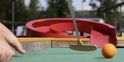 En klubba och en orange boll på en bangolfbana, ett rött hinder rakt fram, en hand som håller i kanten till vänster.