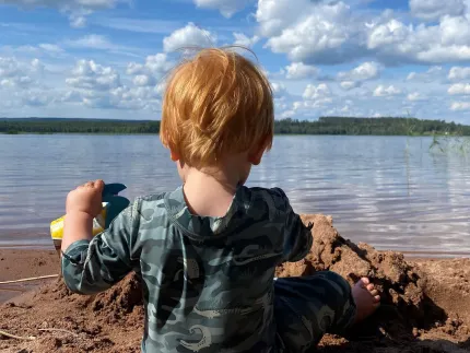 En liten pojke som sitter ner i sanden och har sjön framför sig.